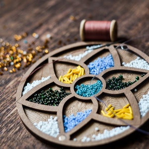 May include: A wooden bead organizer with six compartments filled with different colored beads. The organizer is shaped like a flower with five petals. There is a spool of thread and loose beads scattered on a wooden surface.