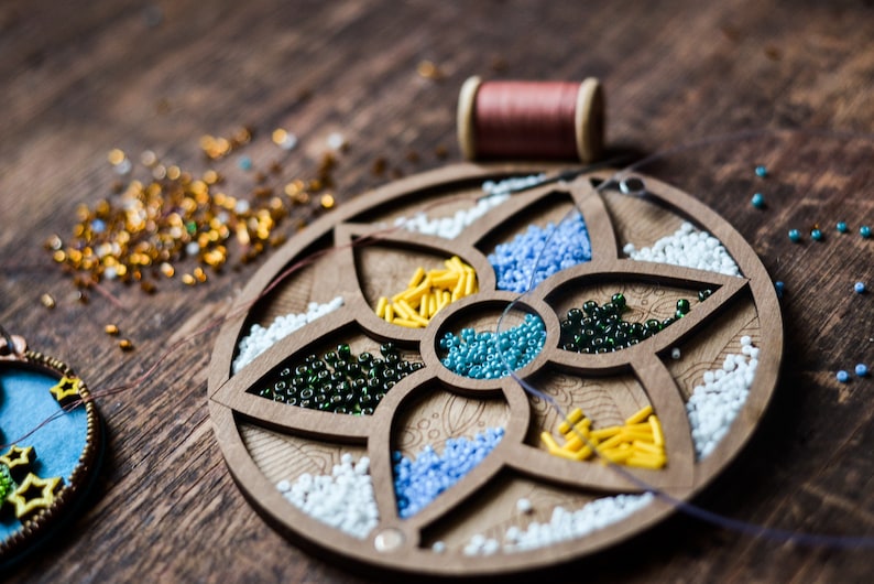 May include: A wooden bead organizer with six compartments filled with different colored beads. The organizer is shaped like a flower with five petals. There is a spool of thread and loose beads scattered on a wooden surface.
