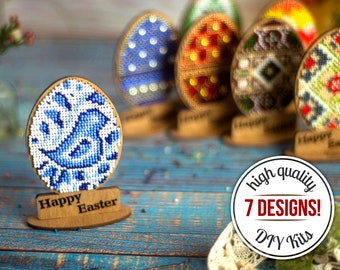 Può includere: Sette diversi design di kit fai-da-te per uova di Pasqua. Ogni kit include un uovo di legno con un design di perline colorate. I design includono un uccello blu, un motivo floreale giallo e arancione, un motivo geometrico rosso e bianco e un motivo geometrico verde e marrone. I kit sono esposti su una superficie di legno blu con il testo "Buona Pasqua" sulla base dell'uovo.