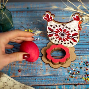 Puede incluir: Un huevo de Pascua rojo se encuentra en un soporte de huevos de madera con forma de flor. El soporte est&aacute; decorado con bordados rojos y blancos. Una figura de pollo con cuentas se encuentra detr&aacute;s del soporte.