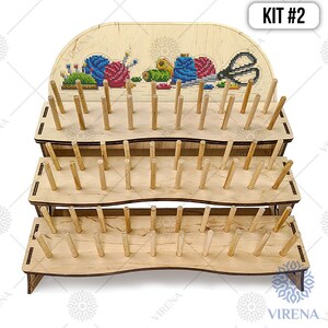 May include: A three-tier wooden stand with dowels for holding embroidery floss. The top tier has a decorative panel with a cross-stitch design of sewing supplies. The stand is labeled "KIT #2".