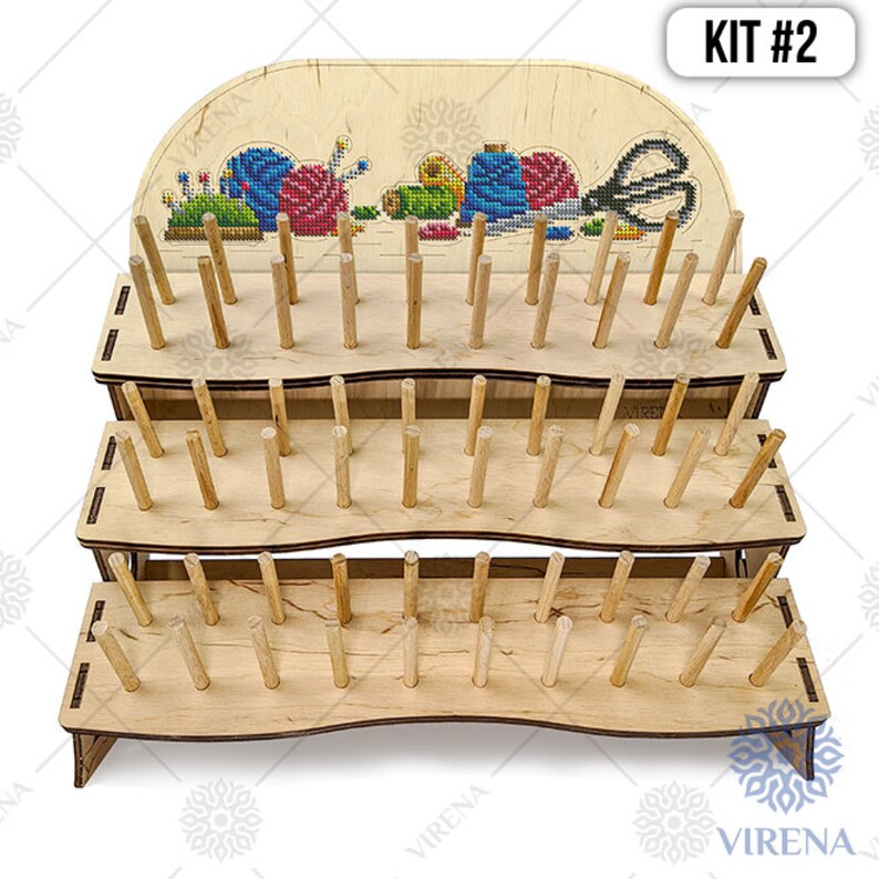 May include: A three-tier wooden stand with dowels for holding embroidery floss. The top tier has a decorative panel with a cross-stitch design of sewing supplies. The stand is labeled "KIT #2".