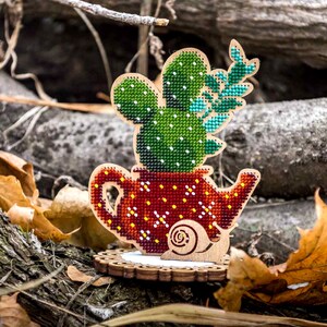 May include: A wooden cactus decoration with a red teapot-shaped pot, adorned with yellow and white dots. The cactus is green with white dots and teal accents. The decoration sits on a wooden base with a snail design.