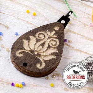 Op de afbeelding: Een houten naaldinrijger met een donkerbruine afwerking en een lichtgekleurd bloemenontwerp. De inrijger heeft een zwarte metalen haak en een klein gaatje. Een ronde sticker leest "wooden needle" en "36 designs threaders".