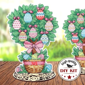 Può includere: Un kit fai-da-te per ricamo a punto croce con un albero di Pasqua colorato con uova e un cestino. Il kit include una base in legno con un design floreale decorativo. Il testo "high quality DIY KIT" è su un cerchio bianco e nero con un uccello.