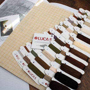 May include: A cross-stitch kit featuring a beige Aida cloth, a colour chart, and a selection of DMC embroidery floss in various shades of brown, beige, and green. The floss is neatly arranged on labelled cards with the text "LUCAS" printed on them.