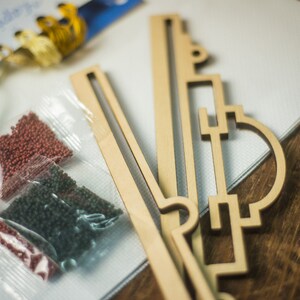 Op de afbeelding: Een close-up van een diamant schilderset met een houten frame, kralen in plastic zakjes en borduurgaren. De kralen zijn rood, groen en bruin. Het frame is gemaakt van lichtbruin hout en heeft een geometrisch ontwerp.