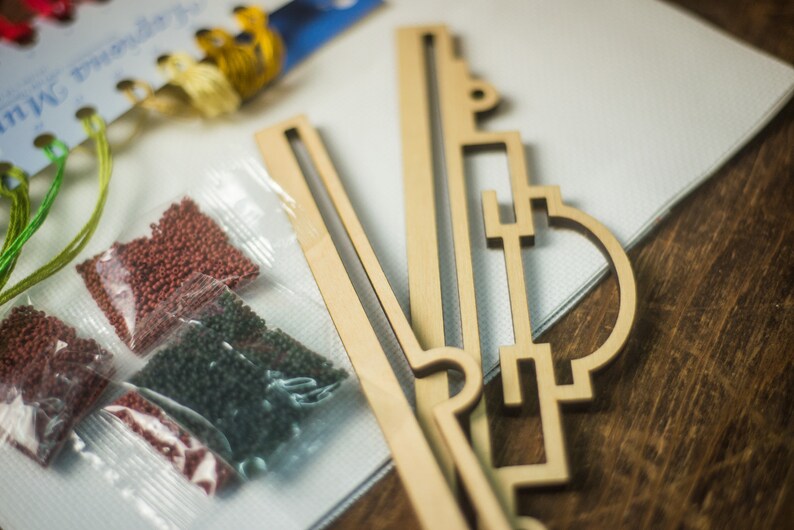 Op de afbeelding: Een close-up van een diamant schilderset met een houten frame, kralen in plastic zakjes en borduurgaren. De kralen zijn rood, groen en bruin. Het frame is gemaakt van lichtbruin hout en heeft een geometrisch ontwerp.