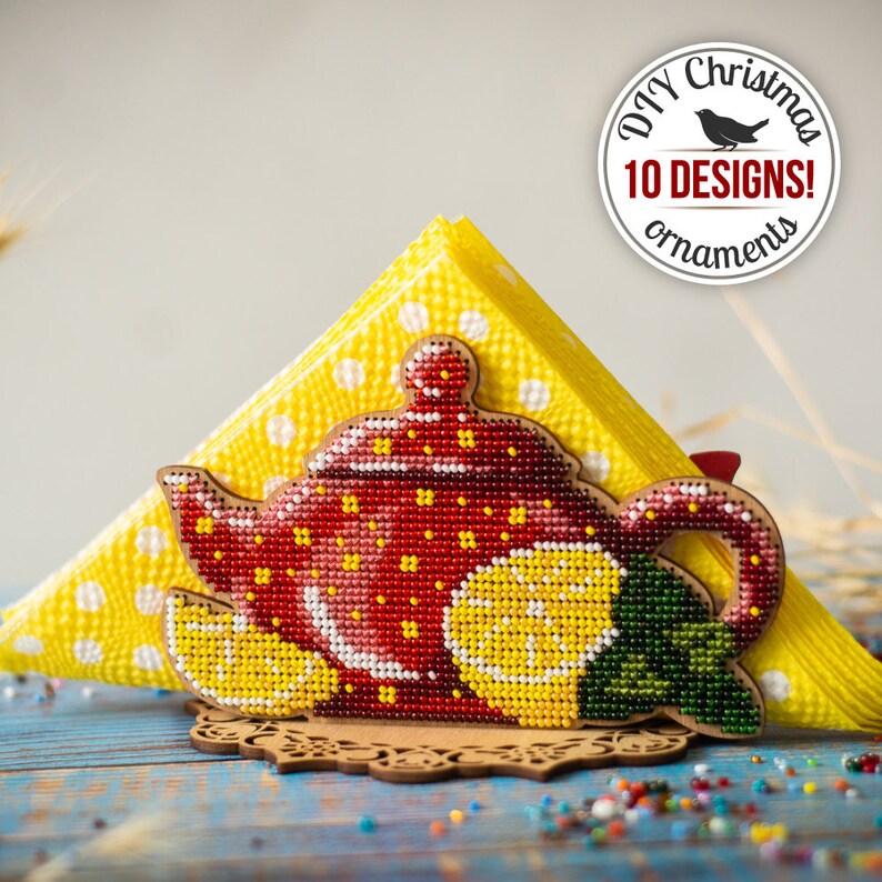 May include: A wooden napkin holder shaped like a teapot with red, yellow, and green beaded details. The holder is decorated with a lemon and a green leaf. The holder is sitting on a blue wooden surface with yellow and white polka dot napkins.