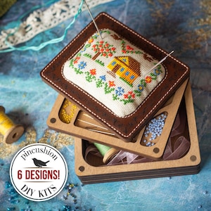 Może przedstawiać: Brązowa poduszka na szpilki w skórzanej ramie z haftowanym krzyżykiem wzorem domu i drzewa. Poduszka na szpilki znajduje się na drewnianym organizerze z przegródkami na koraliki i nici. Obraz zawiera tekst "pincushion 6 DESIGNS! DIY KITS".