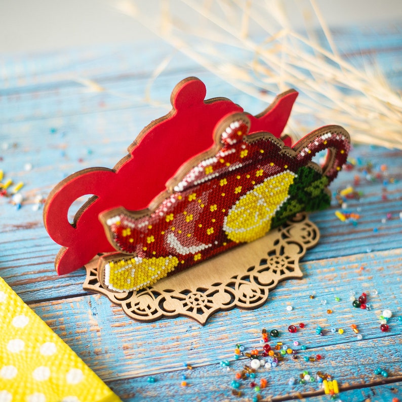 May include: A red wooden napkin holder shaped like a teapot with a beaded design of lemons and flowers. The holder is on a wooden base with a decorative cutout design.