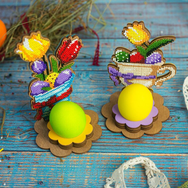 Pu&ograve; includere: Due porta uova di Pasqua in legno decorati con ricami di perline colorate. Un porta uova presenta un mazzo di fiori in un vaso, l'altro una tazza da t&egrave; con fiori. Ogni porta uova contiene un uovo giallo.