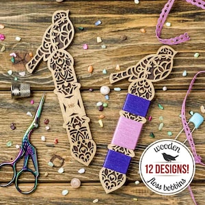 Könnte beinhalten: Holz-Stickgarnspulen mit Vogeldesign. Die Spulen sind hellbraun mit aufwendigen Ausschnitten. Eine Spule enthält rosa und blaues Garn. Eine schillernde Schere und ein Fingerhut sind ebenfalls sichtbar.