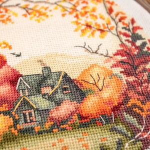 May include: Cross-stitch embroidery of a house with a green roof surrounded by autumn leaves in shades of orange, red, and yellow.