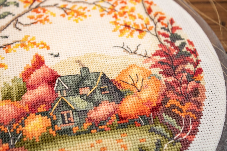 May include: Cross-stitch embroidery of a house with a green roof surrounded by autumn leaves in shades of orange, red, and yellow.