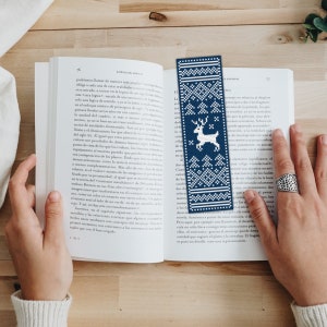 May include: A blue and white knitted bookmark with a reindeer design. The bookmark has a white background with a blue border and a repeating pattern of snowflakes and trees. The reindeer is in the center of the bookmark and is facing to the right.