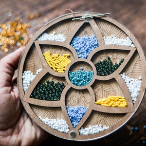 May include: A wooden bead organizer with six compartments, each filled with different colored beads. The organizer is shaped like a flower with a circular base.