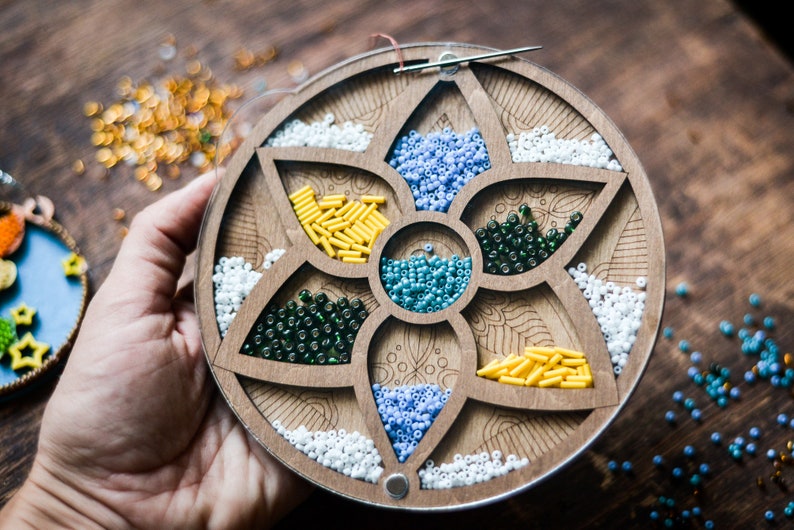 May include: A wooden bead organizer with six compartments, each filled with different colored beads. The organizer is shaped like a flower with a circular base.