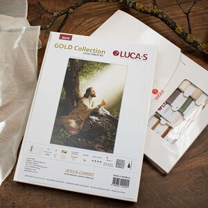 May include: A cross-stitch kit featuring a painting of Jesus Christ praying in a woodland setting. The kit includes white fabric, floss, and instructions. The kit is labelled "GOLD Collection" and "JESUS CHRIST".