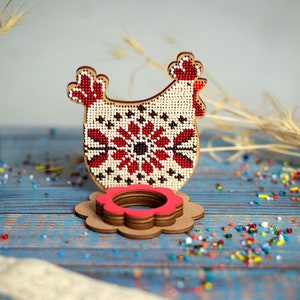 Puede incluir: Un soporte para huevos de Pascua de madera con forma de pollo con un dise&ntilde;o bordado en rojo y blanco. El soporte est&aacute; hecho de madera y tiene una base con forma de flor roja y blanca.