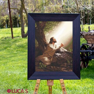 May include: A framed cross-stitch artwork depicting a man kneeling in prayer under a tree with a beam of light shining down on him.
