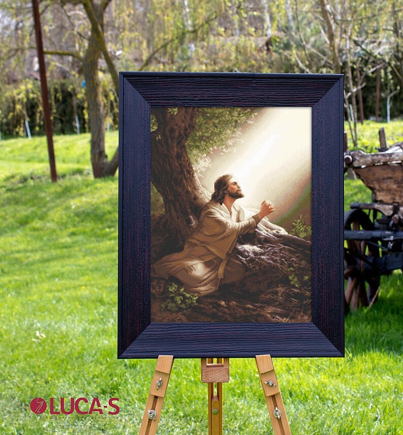 May include: A framed cross-stitch artwork depicting a man kneeling in prayer under a tree with a beam of light shining down on him.