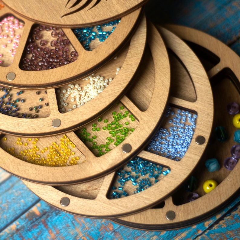 May include: A wooden bead storage organizer with multiple rotating compartments. Each compartment is filled with colorful beads, including pink, brown, white, green, blue, and turquoise.