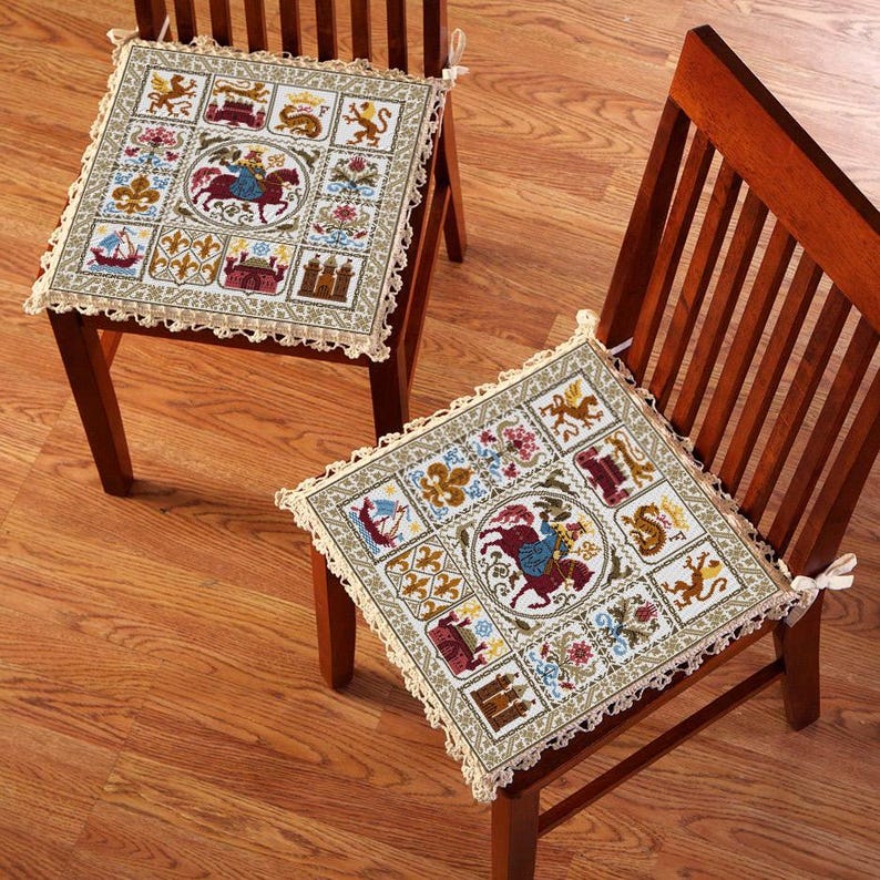 K&ouml;nnte beinhalten: Zwei beige und braune bestickte Stuhlkissen mit einem mittelalterlichen Tapisserie-Design, das einen Ritter zu Pferd, Burgen und andere Symbole zeigt. Die Kissen befinden sich auf Holzst&uuml;hlen mit einer hellen Holzoptik.