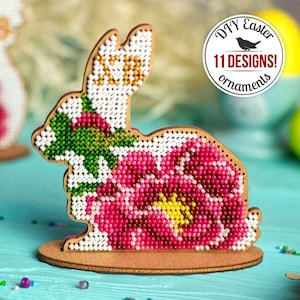 Può includere: Una statuetta di coniglio di legno con un design a fiore rosa, decorata con perline colorate. Il coniglio è in piedi su una base di legno. L'immagine include il testo "DIY Easter 11 DESIGNS! ornaments".