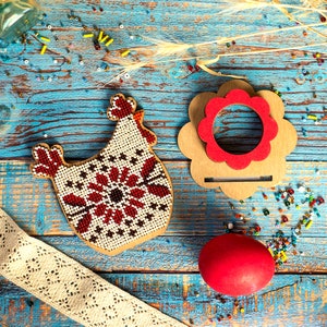 Puede incluir: Una gallina roja de cuentas con fondo blanco y un soporte de madera de forma de flor roja con un centro rojo. La gallina y el soporte est&aacute;n sobre una superficie de madera azul con cuentas rojas y blancas esparcidas alrededor.