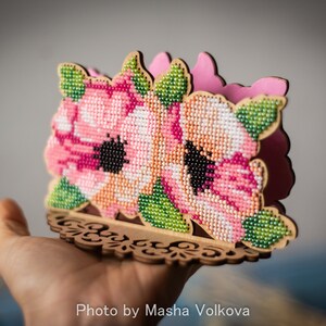 May include: A wooden napkin holder with a floral design. The holder is decorated with pink and white flowers with green leaves, all made of beads. The holder has a decorative, cutout base.