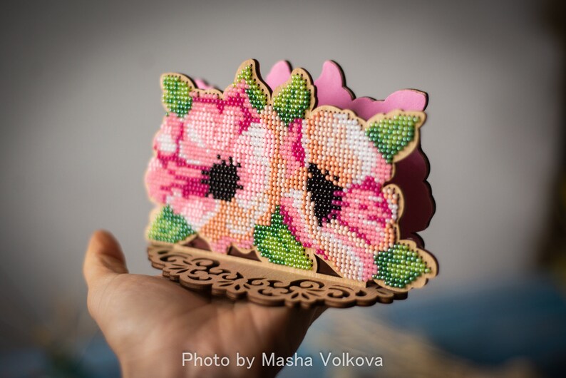 May include: A wooden napkin holder with a floral design. The holder is decorated with pink and white flowers with green leaves, all made of beads. The holder has a decorative, cutout base.