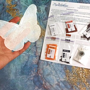 May include: A white butterfly-shaped canvas with a grid pattern for beading. The canvas is surrounded by five small plastic bags containing different colored beads. The bags are labeled with numbers and bead codes. The text on the paper reads "Полезные советы:" which translates to "Helpful tips:"