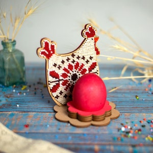 Puede incluir: Un soporte de huevos de Pascua de madera con forma de gallina con cuentas, con un huevo rojo en su interior. La gallina est&aacute; decorada con un patr&oacute;n de cuentas blancas, rojas y marrones.