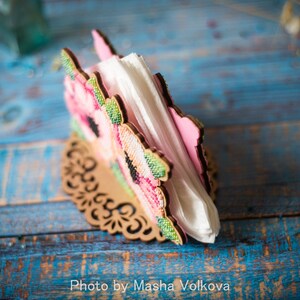 May include: A wooden napkin holder with a floral design featuring pink and green flowers. The holder is on a blue and white wooden surface.