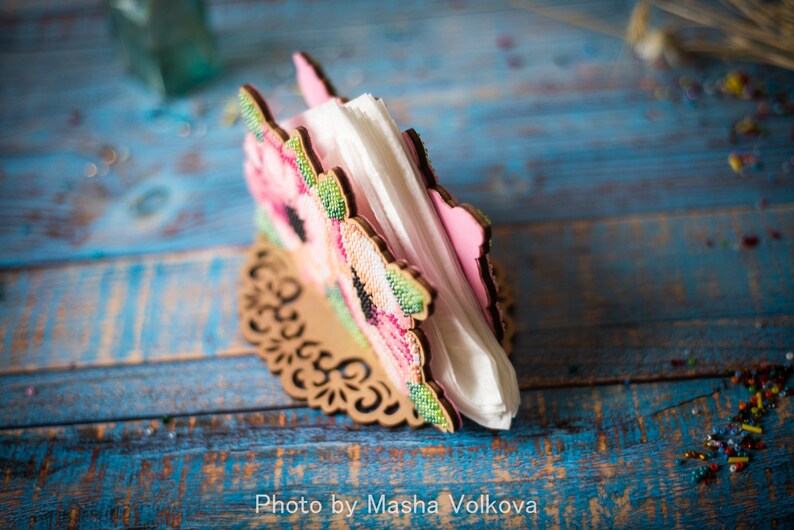 May include: A wooden napkin holder with a floral design featuring pink and green flowers. The holder is on a blue and white wooden surface.