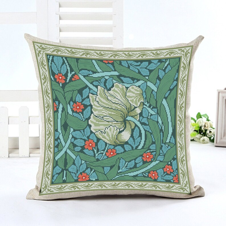 May include: A decorative pillow with a floral design featuring a large white flower with green leaves and orange flowers on a teal background. The pillow has a beige border with a green and white pattern.
