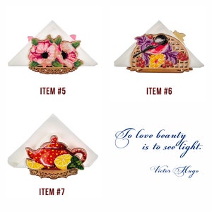 May include: Three wooden napkin holders with embroidered designs. The first holder features pink flowers, the second has a bird and berries, and the third has a red teapot with lemons.