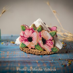 May include: A wooden napkin holder with a floral design. The holder is decorated with pink and green flowers and leaves. The holder is sitting on a blue wooden table.