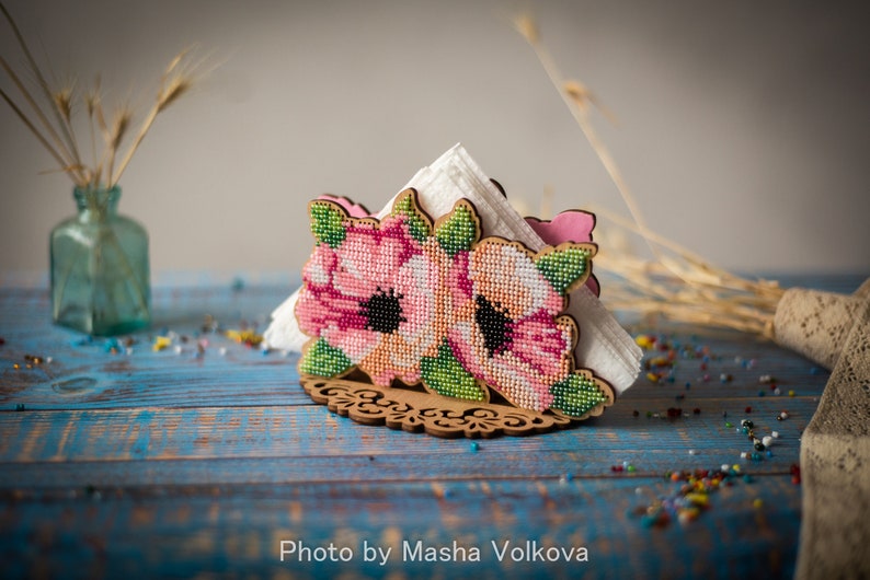 May include: A wooden napkin holder with a floral design. The holder is decorated with pink and green flowers and leaves. The holder is sitting on a blue wooden table.