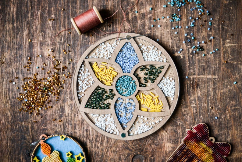 May include: A wooden bead organizer with a flower design. The organizer has seven compartments filled with different colored beads. The beads are in shades of white, blue, green, and yellow.
