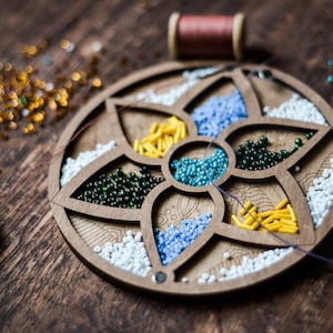 May include: A wooden bead organizer with six compartments filled with different colored beads. The compartments are shaped like flower petals. There are white, blue, green, yellow, and turquoise beads.