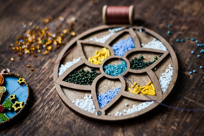 May include: A wooden bead organizer with six compartments filled with different colored beads. The compartments are shaped like flower petals. There are white, blue, green, yellow, and turquoise beads.
