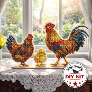 May include: A cross-stitch DIY kit depicting a rooster, hen, and chick. The rooster has a red comb and a blue tail. The hen is brown and the chick is yellow. The kit is displayed on a wooden base, with a window and flowers in the background.