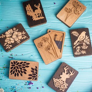 May include: Six wooden boxes with different laser-cut designs. The designs include a deer in a forest, a bird on a branch, a flower, a dandelion, a geometric pattern, and a deer with flowers. The boxes are on a blue wooden surface.