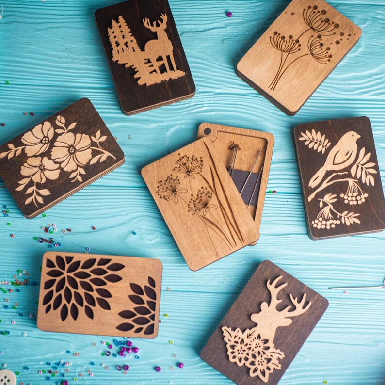 May include: Six wooden boxes with different laser-cut designs. The designs include a deer in a forest, a bird on a branch, a flower, a dandelion, a geometric pattern, and a deer with flowers. The boxes are on a blue wooden surface.