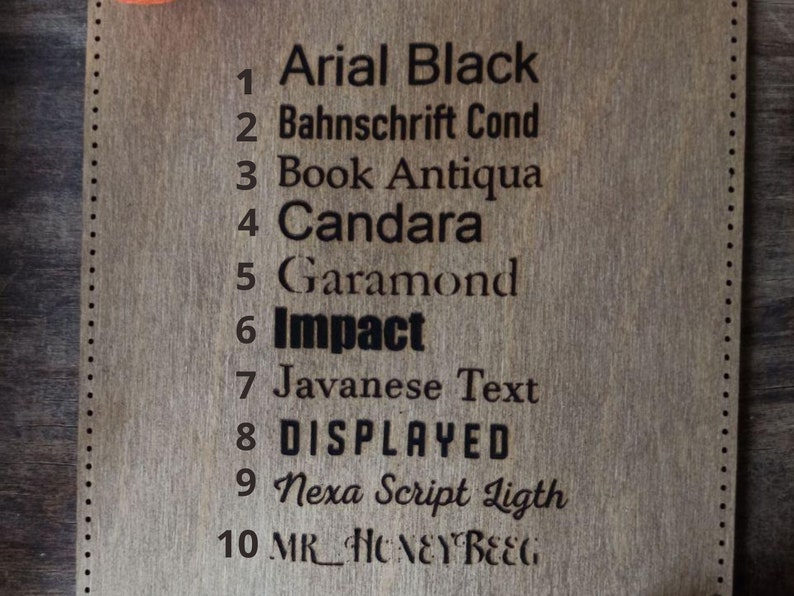 May include: A close-up of a wooden board with a list of 10 different fonts, numbered 1 through 10. The fonts are: Arial Black, Bahnschrift Cond, Book Antiqua, Candara, Garamond, Impact, Javanese Text, DISPLAYED, Nexa Script Ligth, and MR HONEY BEEG.