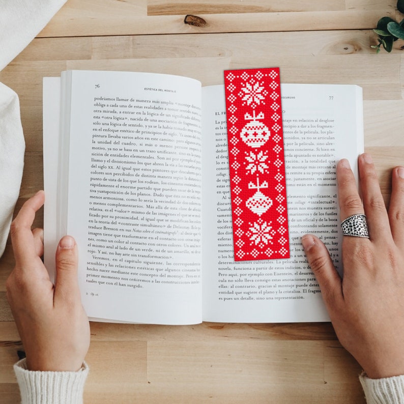 May include: Red and white Christmas-themed bookmark with a snowflake pattern and ornaments. The bookmark is made of fabric and has a fringed edge.