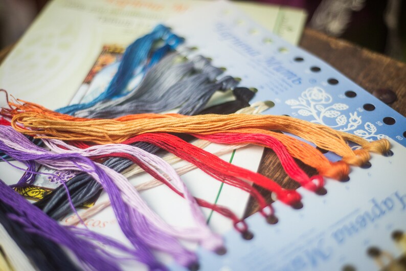 Op de afbeelding: Een close-up van een verscheidenheid aan borduurgaren spoelen in verschillende kleuren, waaronder paars, oranje, rood en blauw. Het garen is in een waaier vorm op een wit oppervlak met een blauw en wit bedrukte kaart gerangschikt.