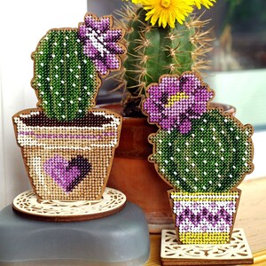 May include: Two wooden cactus decorations with beaded details. One is tall with a purple flower, the other is round. Both are in pots with heart and floral designs. They sit on decorative wooden coasters, with a real cactus in the background.